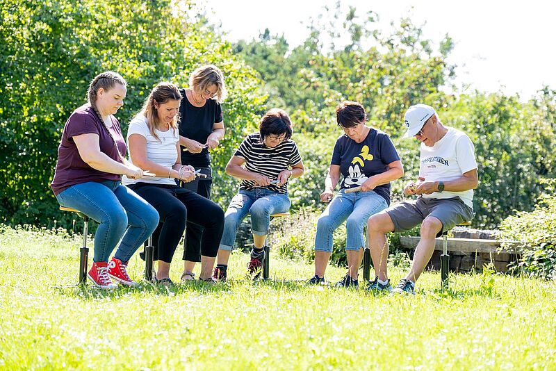 Personengruppe sitzt auf sonniger Wiese und schnitzt Grünholz