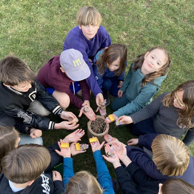 Kindergruppe auf einer Wiese steht um ein Insektenhotel und hält Bienenmodelle auf den Händen