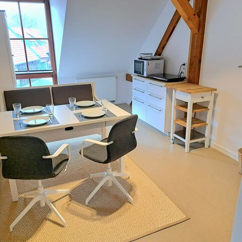 Blick auf eine Sitz-/Essecke mit grau-weißen Stühlen, weißem Tisch und Sitzbank in einem Fachwerkraum mit Kochnische im Hintergrund