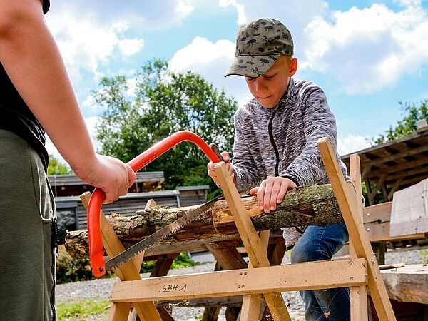 Junge mit Basecap sägt unter Anleitung konzentriert ein Holzstück durch
