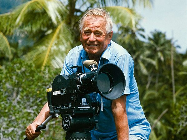 Heinz Sielmann steht lächelnd in einem kurzärmeligen Hemd an der Kamera, hinter ihm eine tropische Landschaft
