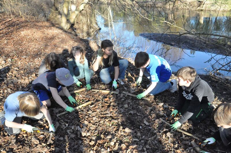 Eine Gruppe von Kindern hockt im Wald an einem Bachlauf und sägt Äste