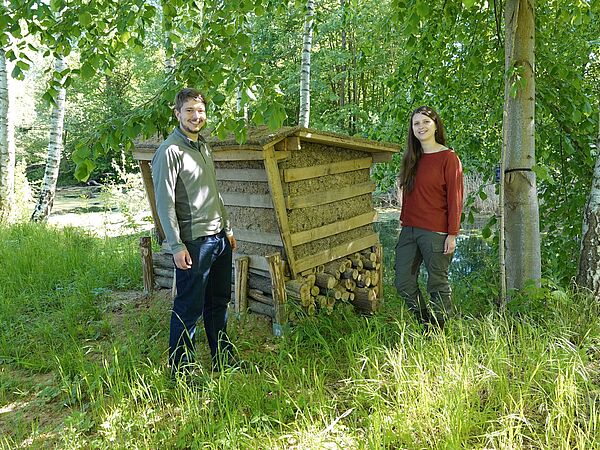 Junger Mann und Frau stehen von zwei Seiten neben einer Nisthilfe aus Holz