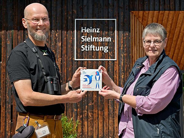 Zwei Personen stehen mit einer Auszeichnung für ein schwalbenfreundliches Haus in den Händen vor einer Holzwand