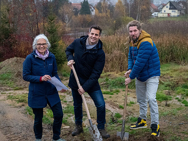 Spatenstich Oberviechtach Drei Personen stehen im Frühwinter auf einer Brache. Links steht eine Frau mit dickem blauen Mantel und grauen Haaren und hält ein Dokument in den Händen. In der Mitte hält ein Mann im Wintermantel einen Spaten auf den er einen Fuß gestellt hat. Rechts steht ein weiterer Mann in einer Daunenjacke und hält ebenfalls einen Spaten.